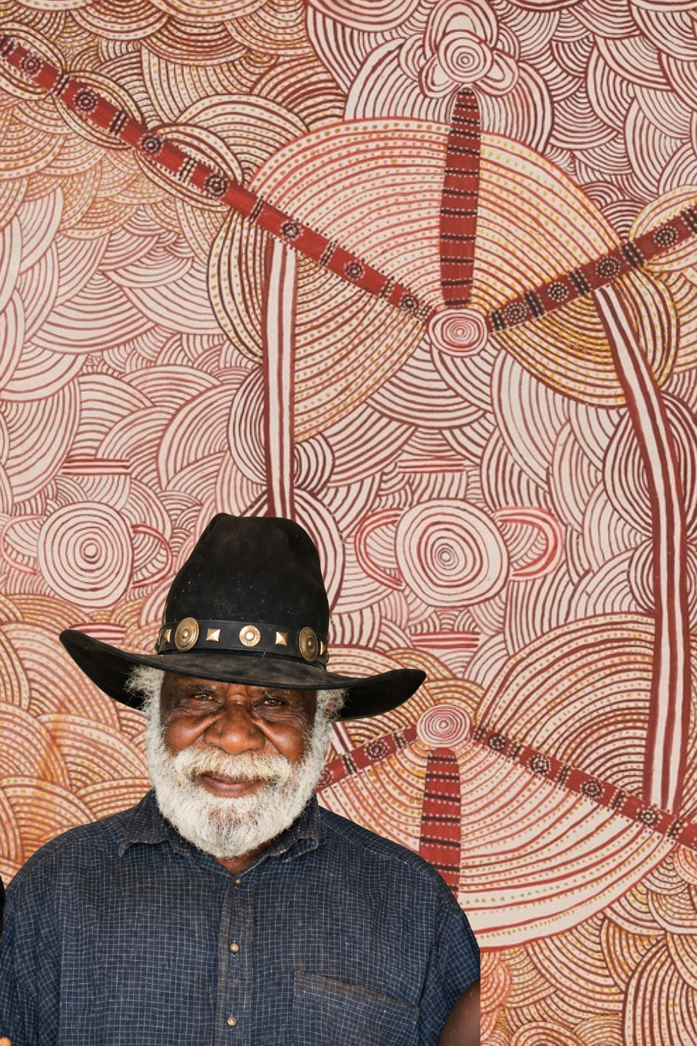 Long Jack Phillipus Tjakamarra – Papunya Artist Long Jack Phillipus Tjakamarra, pioneering Papunya artist, pictured against a Western Desert painting featuring traditional geometric designs.