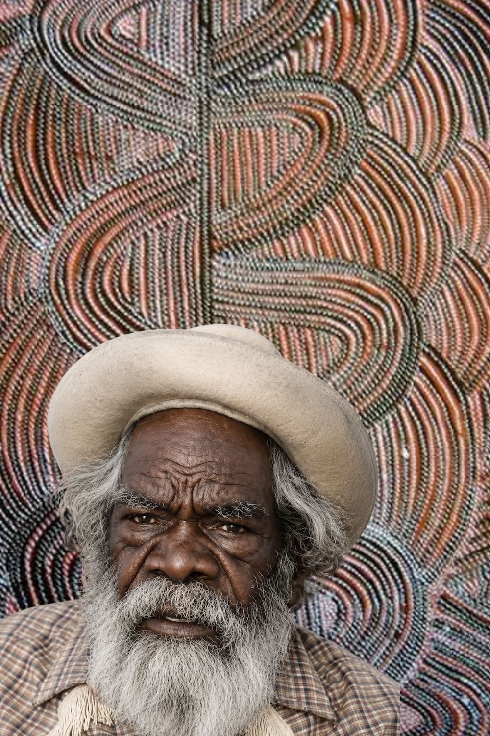 Portrait of Mick Namarari Tjapaltjarri wearing a pale hat, with white hair and beard, in front of a Western Desert painting featuring interlocking curved lines and fine dotting in earthy colours.