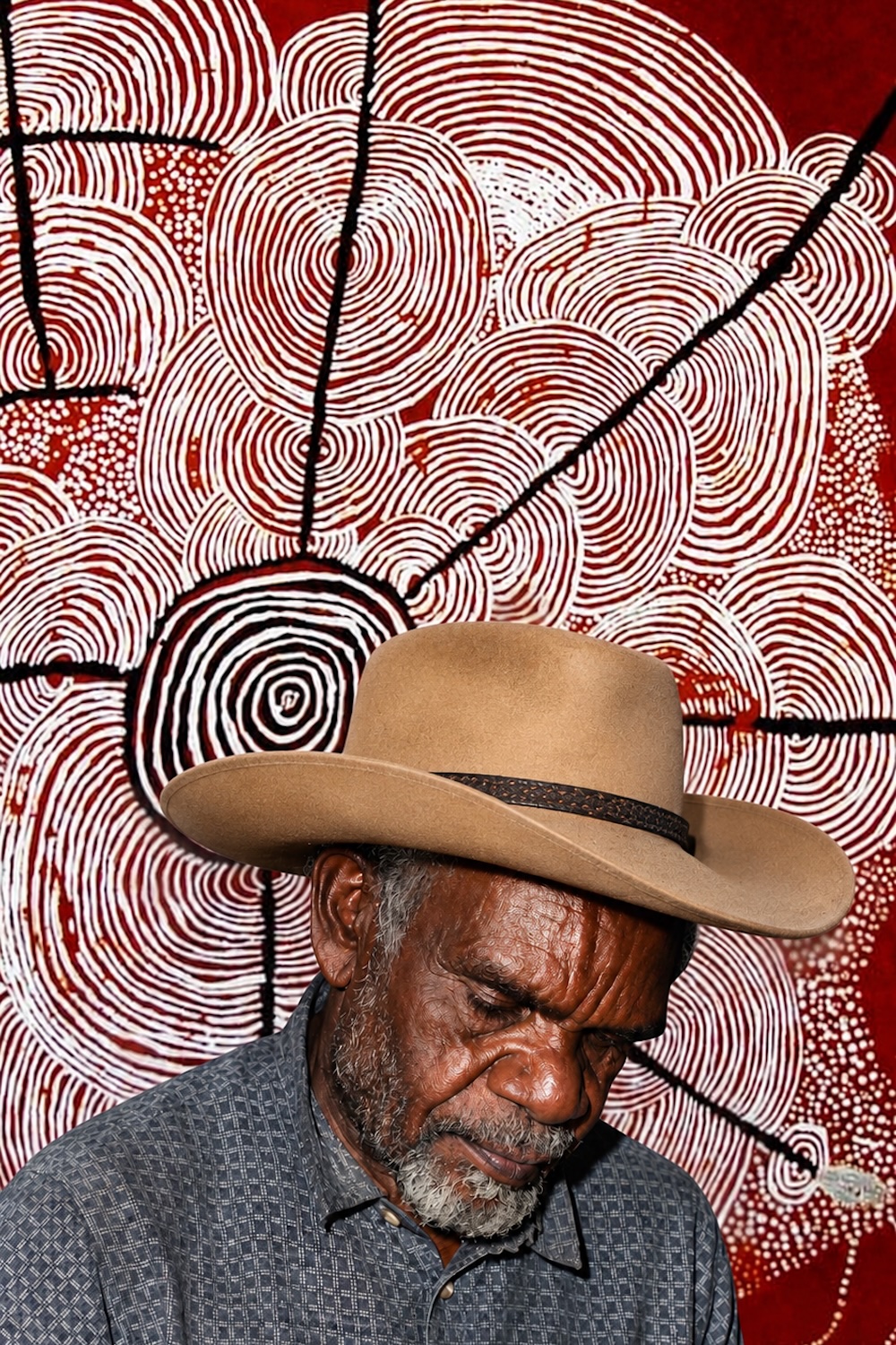 Portrait of Shorty Lungkata Tjungurrayi wearing a tan hat, in front of a red Western Desert painting featuring concentric circles, white curved lines and intricate dotting.