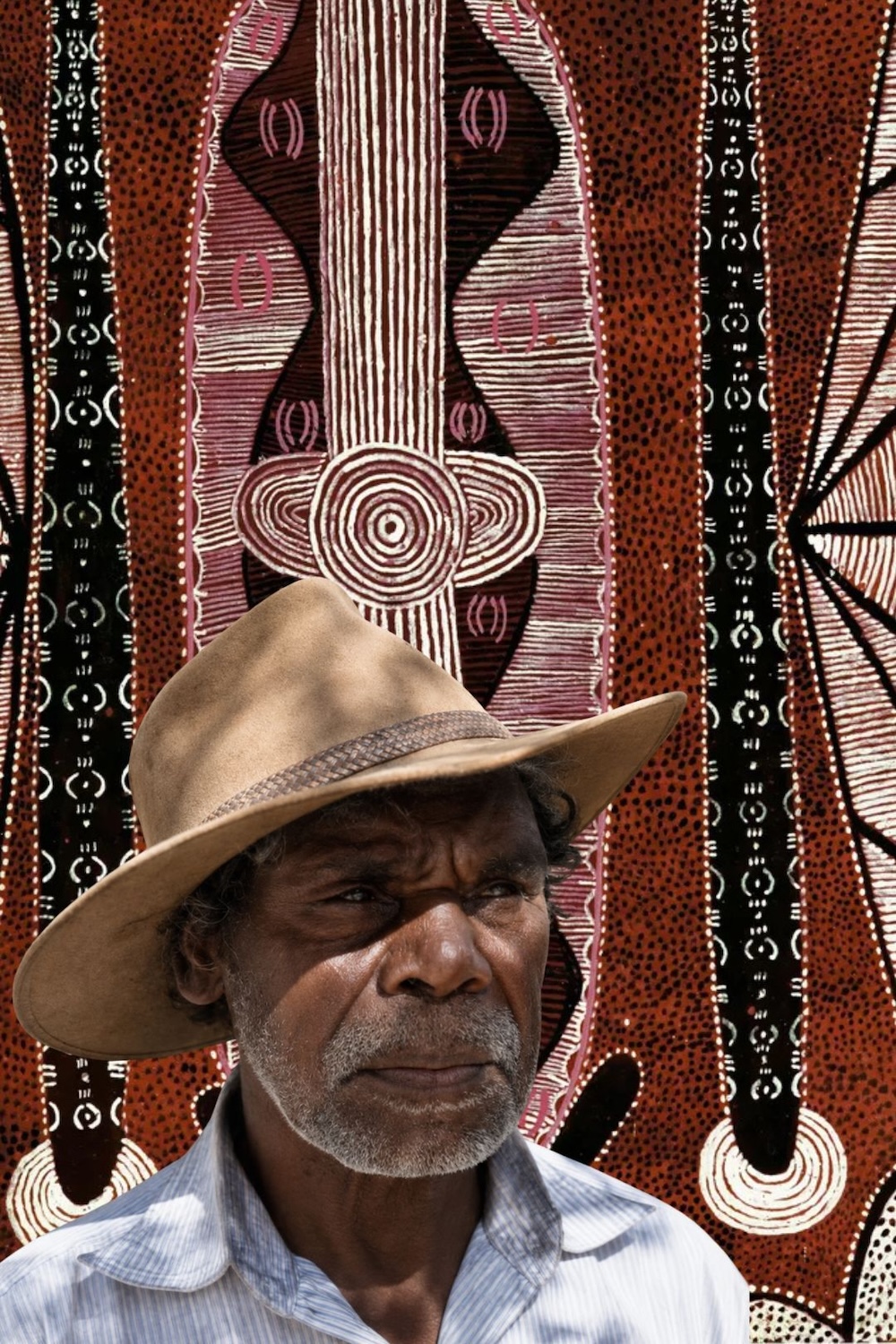 Portrait of Tim Leura Tjapaltjarri wearing a broad-brimmed hat, in front of a Western Desert painting featuring concentric circles, elongated geometric forms and intricate white markings on a red ochre background.