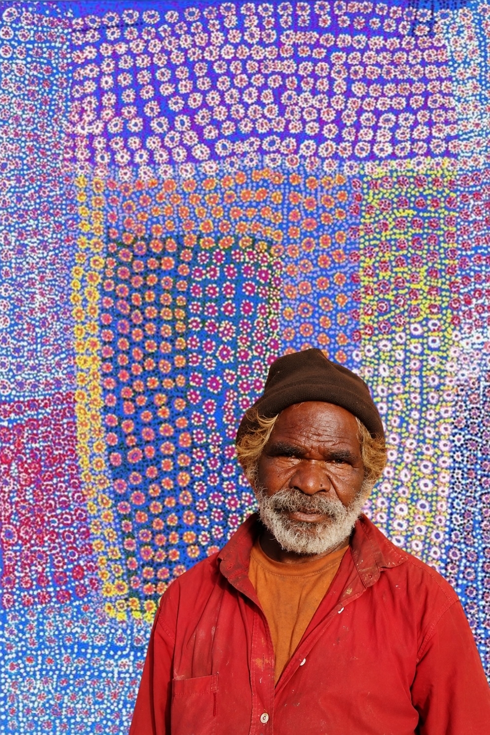 Tommy Mitchell – Ngaanyatjarra Western Desert Artist Portrait of Tommy Mitchell, Ngaanyatjarra Aboriginal artist, standing before a vibrant Western Desert dot painting.