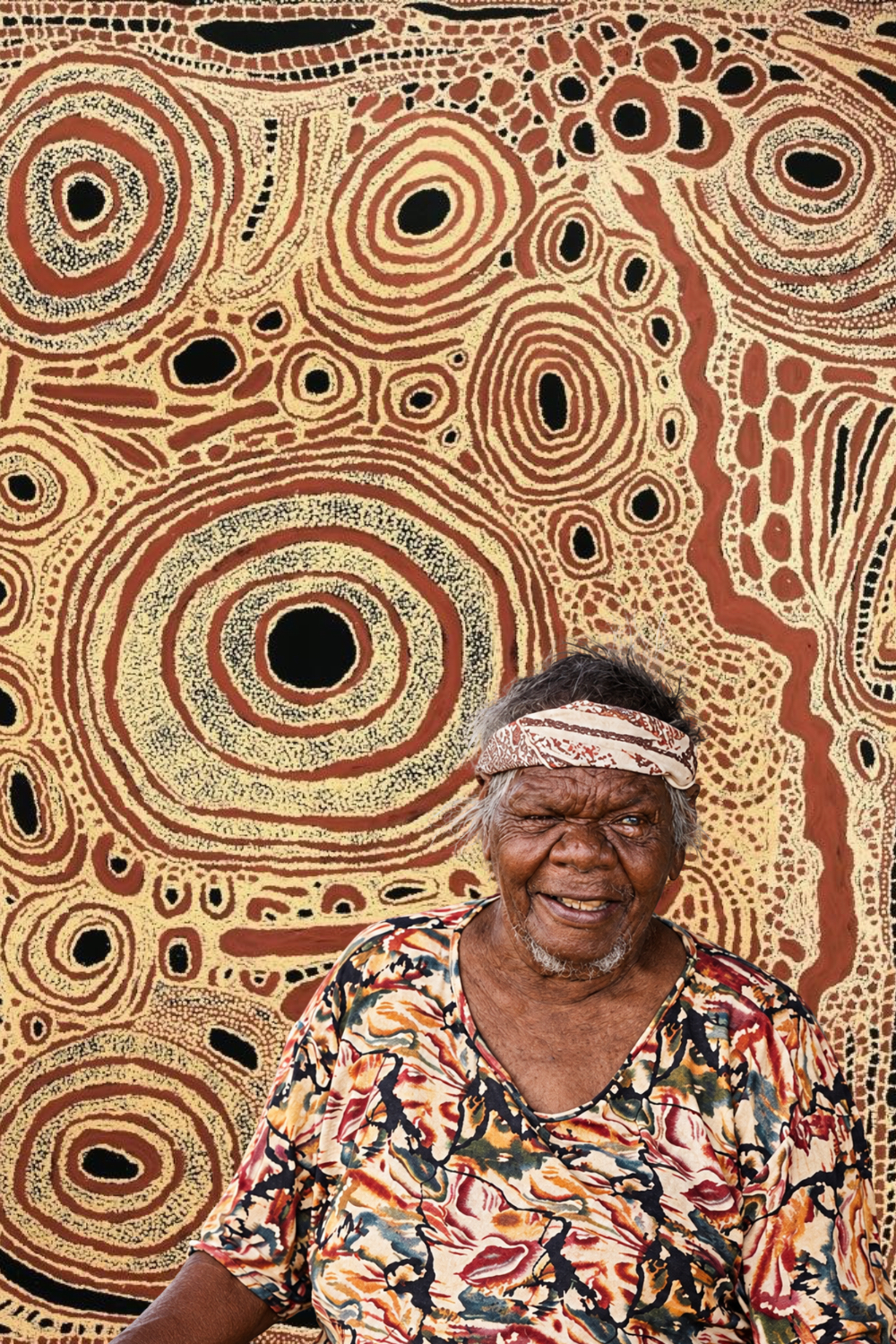 Nyurapayia Nampitjinpa (Mrs Bennett) seated in front of an Aboriginal painting featuring concentric circles in red ochre, cream and black.