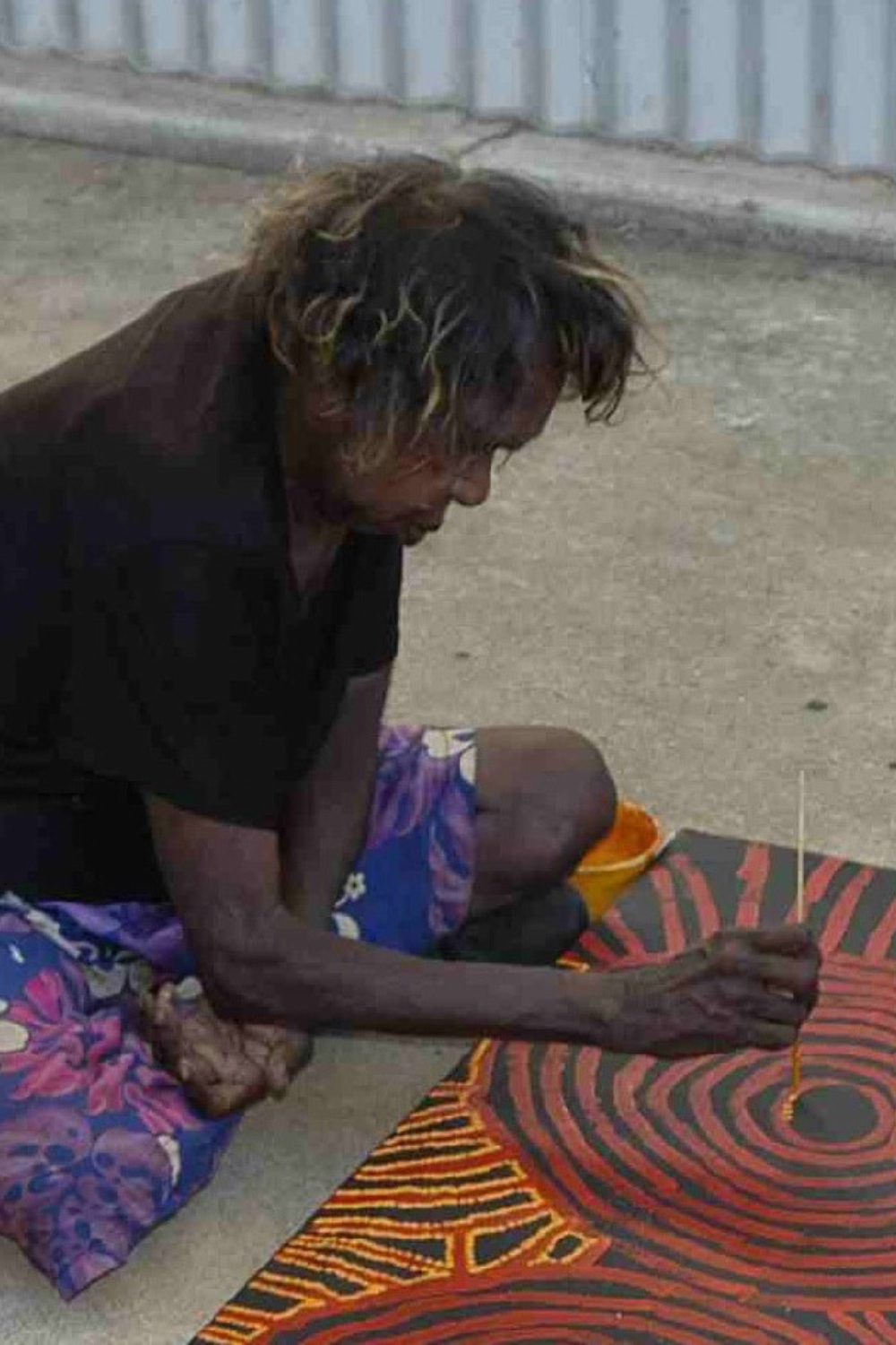 Walangkura Napanangka seated on the ground painting intricate red and orange concentric circles and linear designs on a black canvas.