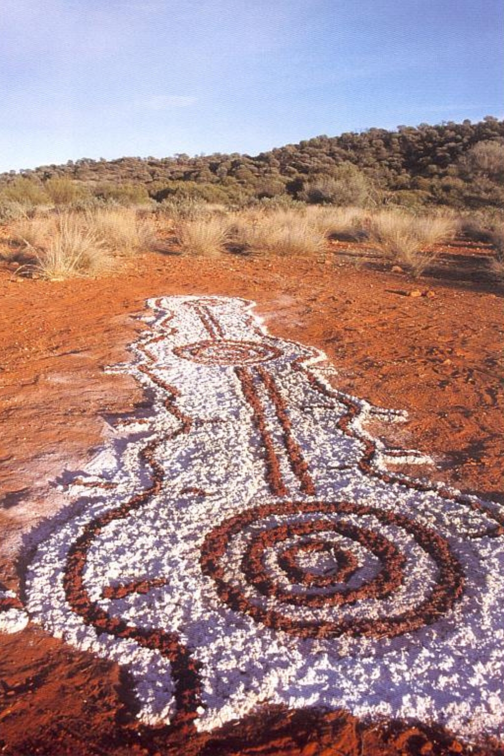 Aboriginal ceremonial sand painting on red desert earth, featuring concentric circles, winding ochre lines and white ground decoration.