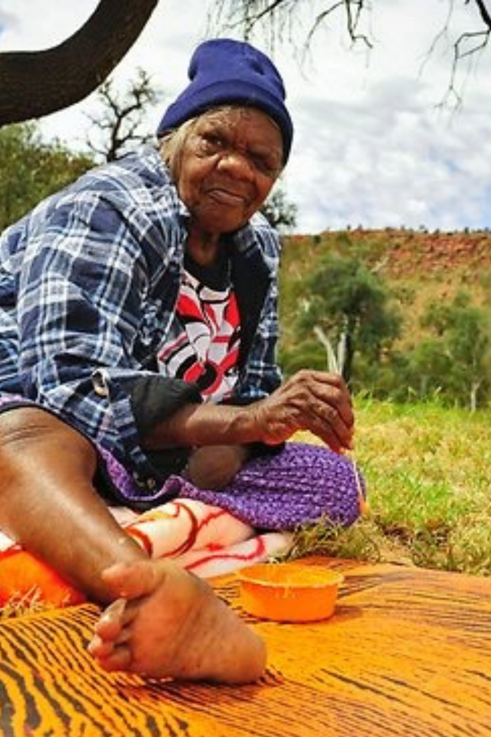 Nyurapayia Nampitjinpa (Mrs Bennett) working on an Aboriginal artwork outdoors, seated in the Western Desert landscape wearing a blue beanie and checked shirt.